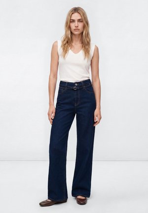 Frau mit langen blonden Haaren, die ein ärmelloses weißes Top, hoch taillierte dunkelblaue Jeans mit weitem Bein und braune flache Schuhe trägt, steht vor einem einfarbigen Hintergrund.