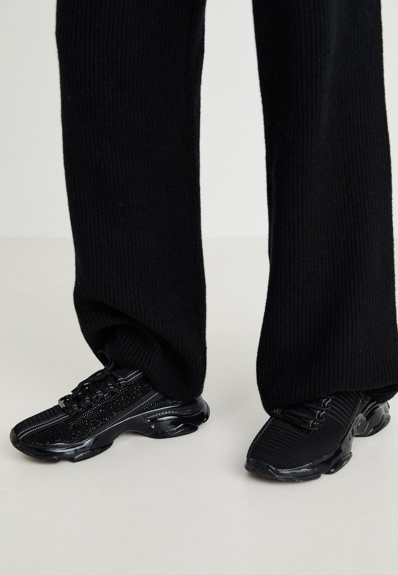 Black sneakers with a textured knit upper, sleek shape, and glossy black sole, featuring subtle reflective accents and lace-up design.