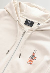 Sweat à capuche zippé crème avec cordons, présentant un graphisme vintage de bouteille Pepsi-Cola et une étiquette de col noire affichant la marque Pepsi x NEXT.