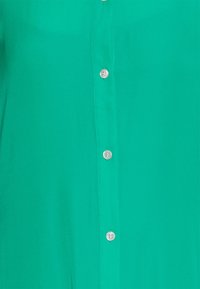 Grön skjorta med knappar gjord av mjukt tyg, som har en rad av fyra vita knappar framtill och en enfärgad yta utan mönster.