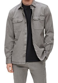 Lichtgrijs overhemd met een klassieke kraag, twee borstzakken en lange mouwen. Gedragen over een zwart T-shirt, gemaakt van gladde stof.