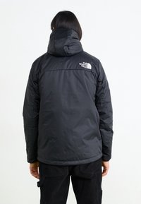 Černá zateplená bunda s kapucí, která má lesklou texturu, prošívané detaily a bílé logo na levém rameni.