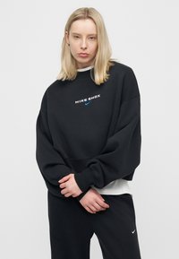 Νεαρή γυναίκα με ίσια ξανθά μαλλιά, που φοράει μαύρο φούτερ Nike Shox και μαύρο παντελόνι, στεκόμενη με τα χέρια μαζεμένα.