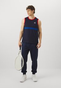 Mornarsko modra športna majica brez rokavov z rdečimi poudarki in modrimi črtami, v kombinaciji z mornarsko modrimi trenirka hlačami, ki drži teniško lopar.