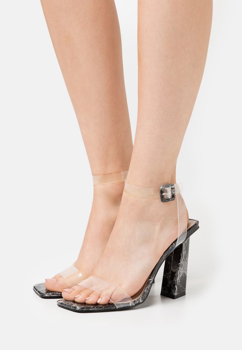 Pieds portant des sandales à talons blocs motif marbre noir avec des brides transparentes et des brides de cheville maintenues par des boucles argentées.