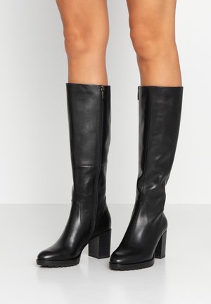 Zwarte leren laarzen met hakken en ritssluitingen aan de zijkant, gedragen op blote benen tegen een effen witte achtergrond.