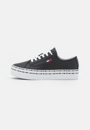 Baskets basses noires avec des lacets blancs, semelle épaisse blanche arborant un motif carré noir, et petit logo rouge, blanc et bleu sur le côté.