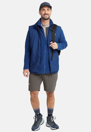 Veste polaire bleue sur un haut gris, associée à un short gris. Sac à dos noir, bottes de randonnée bleues et chaussettes à motifs complètent la tenue.