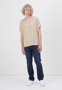 Beige bomull t-skjorte med "GUESS" logo i hvitt, kombineres med mørkeblå jeans og hvite sneakers. Modellen står foran en nøytral bakgrunn.