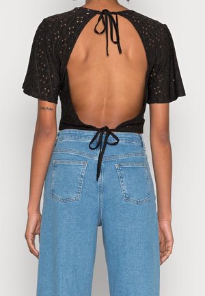 Woman wearing black open-back top with tie closures and high-waisted light blue jeans, shown from the back against a plain background.