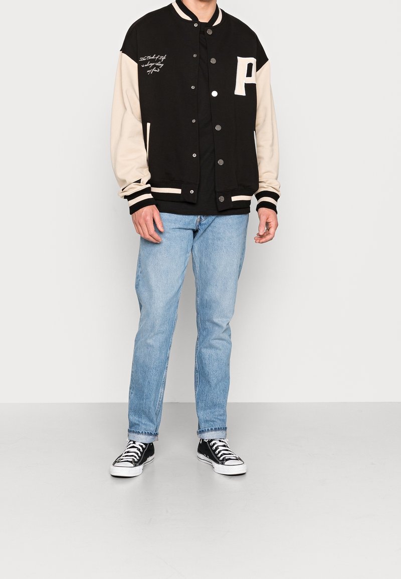Homme portant une veste universitaire noire et beige, un jean bleu clair et des baskets basses noires, debout devant un fond uni.
