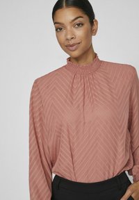 Blus i mjuk rosa med ett strukturerat, zickzackmönster. Har en hög, smockad halsringning och långa ärmar. Kombineras med svarta byxor.