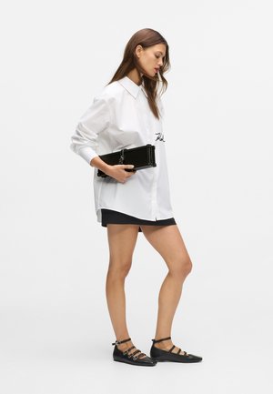 Biała koszula z długim rękawem i czarnymi haftowanymi detalami, zestawiona z czarną dopasowaną spódnicą i czarnymi baletkami na cienkich paskach.