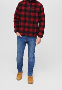 Muž v červené a černé kostkované fleecové bundě, modrých džínách a béžových šněrovacích boots, stojící s jednou rukou v kapse.