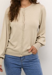 Béžový pletený svetr cardigan s kulatým výstřihem, zapínáním na knoflíky a dlouhými rukávy s žebrovanými manžetami. Kombinováno s modrými džínami.