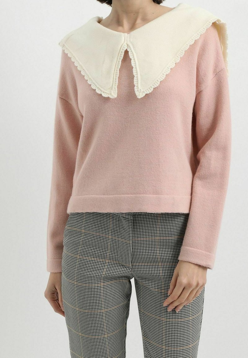 Mujer con un suéter de manga larga rosa y un collar blanco sobredimensionado, llevando pantalones a cuadros en blanco y negro, de pie contra un fondo sencillo.