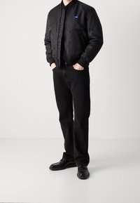 Schwarze Bomberjacke mit gerippten Bündchen und Kragen, kombiniert mit schwarzen Jeans und schwarzen Schuhen. Auffälliges blaues Logo-Patch auf der Jacke.