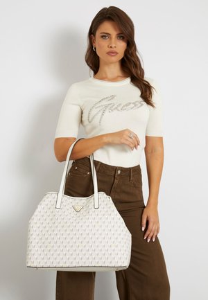 Sac à main blanc avec un motif texturé et un logo, présentant des accents beiges et des poignées doubles. Le modèle porte un haut crème et un pantalon marron.