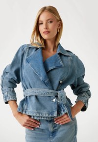 Giacca di jeans azzurra chiara con rever oversize, chiusura a bottoni, vita con cintura e maniche arrotolate. Texture liscia con dettagli a contrasto.