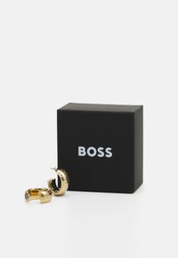 Pendientes de aro dorados con acentos texturizados, acompañados de una caja negra con la marca blanca "BOSS". Superficie lisa, forma circular.