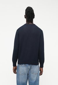 Tumsas zila adīta džempera ar apaļu kakla izgriezumu, ribveida aprocēm un apakšu, ar vaļīgu piegriezumu. Valkāts kopā ar gaiši zilām džinsu biksēm, izceļot ikdienišķu stilu.