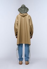 Pardesiu impermeabil kaki cu glugă și croială lejeră, asortat cu blugi albastru deschis cu accente legate. Ținuta este completată de pantofi maro.