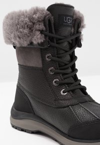 Bota negra impermeable que presenta materiales de cuero y ante, acabado texturizado, cuello de pelo sintético gris, ojales de metal y cordones negros.