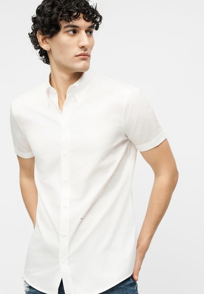 Jeune homme aux cheveux bouclés portant une chemise blanche à manches courtes, regardant sur le côté, les mains derrière le dos.