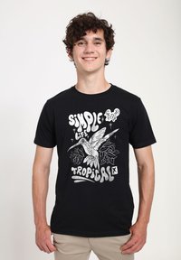 Černé tričko s grafickým designem, na kterém je kolibřík a tropické listy, s bílým obrysem textu "Jednoduchý život tropický."
