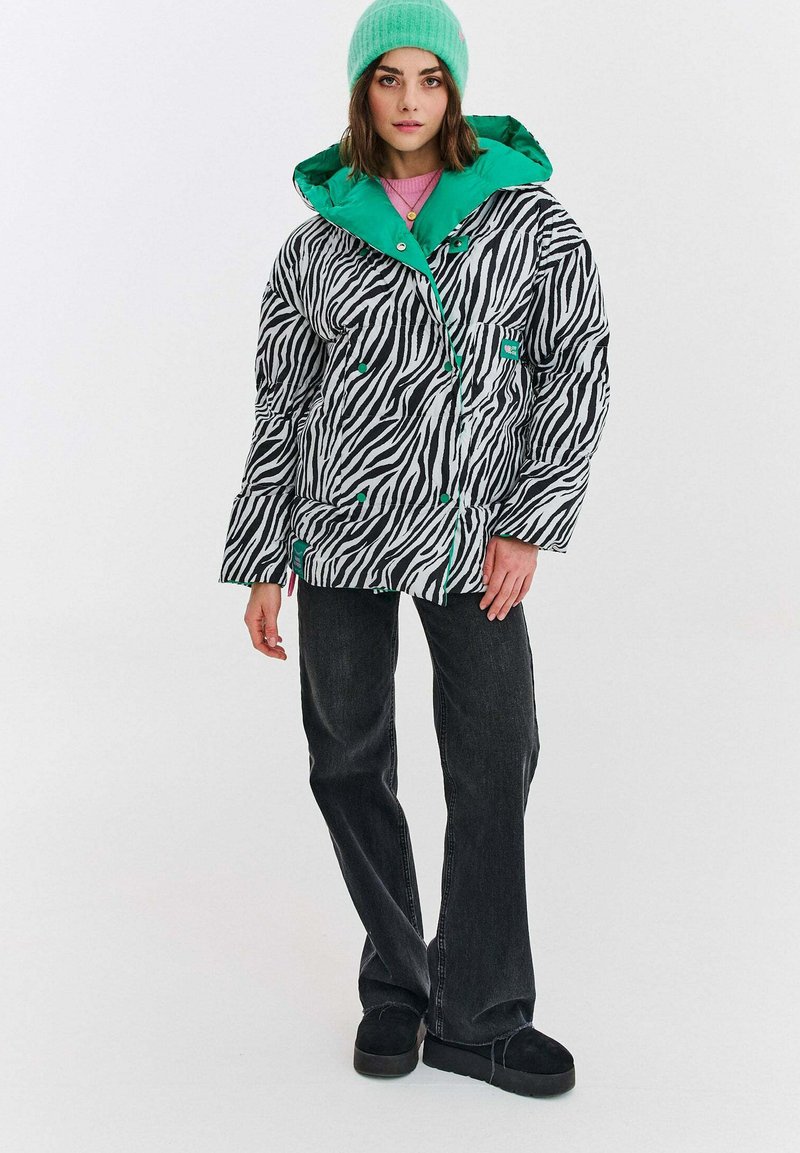 Prošívaná bunda se zebřím vzorem v černé a bílé barvě se zelenou kapucí, doplněná tmavými džíny s rozšířenými nohavicemi a černou obuví. Viditelné zdobené knoflíky.