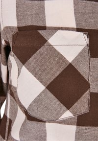 Poche de chemise à carreaux marron et blanc avec des coutures visibles sur un tissu à grand motif à carreaux.