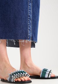 Chanclas coloridas con un diseño de volantes en capas en verde, azul y blanco, montadas sobre una suela plana negra. Estilo casual y de punta abierta.