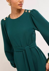 Zelené šaty s dlhými rukávmi, zdobené zlatými gombíkmi v tvare ruží na ramenách, so zaväzovaním v páse a hladkou textúrou látky.