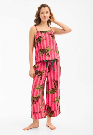 Vrouw staat blootsvoets en draagt een roze gestreept pyjamaset met tijger- en groene plantafdrukken, met één hand die haar haar aanraakt, neutrale achtergrond.