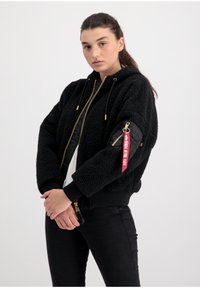 Svart bomberjacka med en texturerad, fluffig utsida, dragkedja fram och ribbade manschetter. Har en sidoficka med en röd etikett.