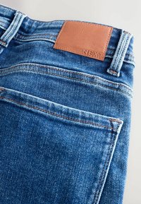 Blugi din denim de culoare albastru mediu, cu un finisaj texturat, având o etichetă din piele maro la talie, embosată cu cuvântul "NEXT" și detalii de cusătură vizibile.