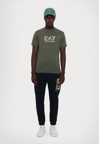 Olivengrønn t-skjorte med korte ermer og logo i gulltekst, kombinert med marineblå joggebukser med hvit logo-print. Brukt med hvite sneakers og en grønn caps.