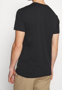 Muž v jednoduchém černém tričku s krátkým rukávem a béžových kalhotách, zobrazený ze zad na jednobarevném bílém pozadí.