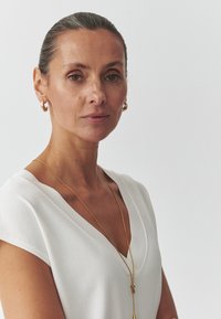 Top bianco a maniche corte con scollatura a V, abbinato a una lunga collana dorata e orecchini a cerchio. La pelle appare liscia, con trucco minimal.