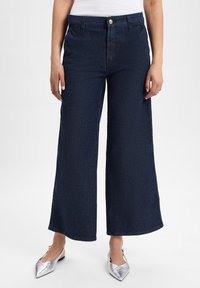 Brede, blauwe denim broek met een hoge taille, voorzien van een knoopsluiting en subtiele textuur. Gecombineerd met metallic zilveren puntige schoenen.