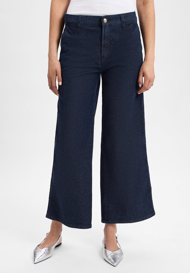 Brede, blauwe denim broek met een hoge taille, voorzien van een knoopsluiting en subtiele textuur. Gecombineerd met metallic zilveren puntige schoenen.