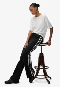 Camiseta blanca de gran tamaño y pantalones negros con rayas laterales blancas. Modelo sentado en un taburete de madera. Zapatos oscuros completan el conjunto.