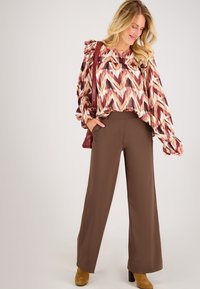 Lange mouwen, vloeiende blouse met een rood en beige zigzagpatroon; gecombineerd met hoge, bruine wijde broek en tan blokhakken laarzen.