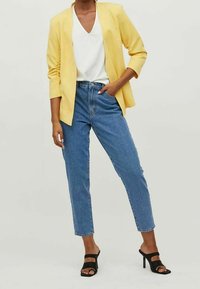 Keltainen bleiseri, jossa on kolme neljäsosaa hihoista, valkoinen V-kaula-aukkoinen toppi, vaaleansiniset korkeavyötäröiset farkut ja mustat nauhalla varustetut korot.