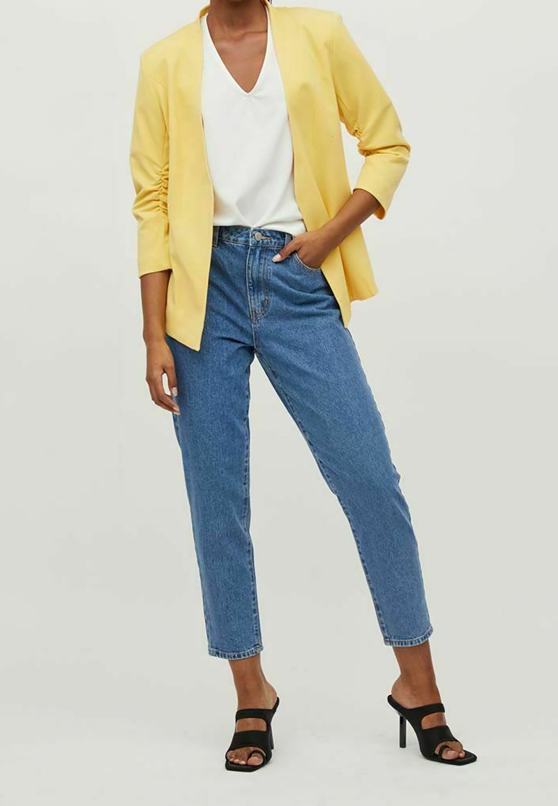 Keltainen bleiseri, jossa on kolme neljäsosaa hihoista, valkoinen V-kaula-aukkoinen toppi, vaaleansiniset korkeavyötäröiset farkut ja mustat nauhalla varustetut korot.