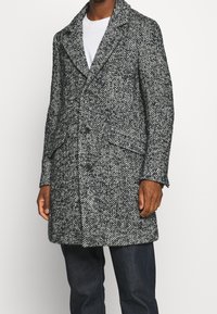 Cappotto grigio con motivo a spina di pesce, caratterizzato da un colletto a revers, chiusura con bottoni e due tasche frontali. Realizzato in tessuto strutturato.