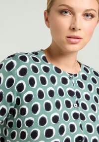 Kobieta w zielonej bluzce z guzikami, w czarno-białym okrągłym wzorze, ukazująca górną część swojej twarzy i tors.