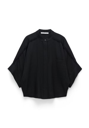 Schwarzes übergroßes Hemd mit Stehkragen, aufgesetzter Tasche vorne und Fransenbesatz entlang der Raglanärmel und Schultern, am Hals mit Knopf verschlossen.