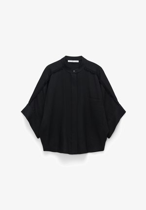 Schwarzes übergroßes Hemd mit Stehkragen, aufgesetzter Tasche vorne und Fransenbesatz entlang der Raglanärmel und Schultern, am Hals mit Knopf verschlossen.