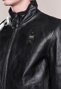 Veste en cuir noir avec un col rabattu, fermeture éclair à l'avant et une finition texturée. Présente une étiquette et un détail à bouton-pression sur la poitrine.